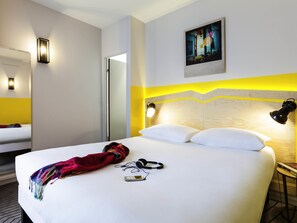 A hotel room with a bed, a bedside table, a lamp, a painting on the wall, and a door leading to another room. - ibis Styles Paris Nation Porte De Montreuil (Paris)