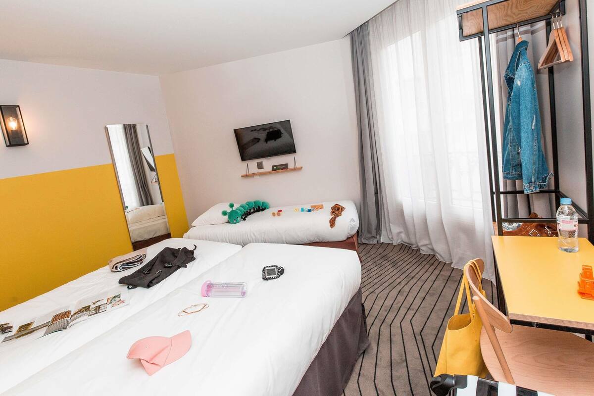 A hotel room with two beds, a TV, a mirror, a table with a chair, and a coat rack with clothes.