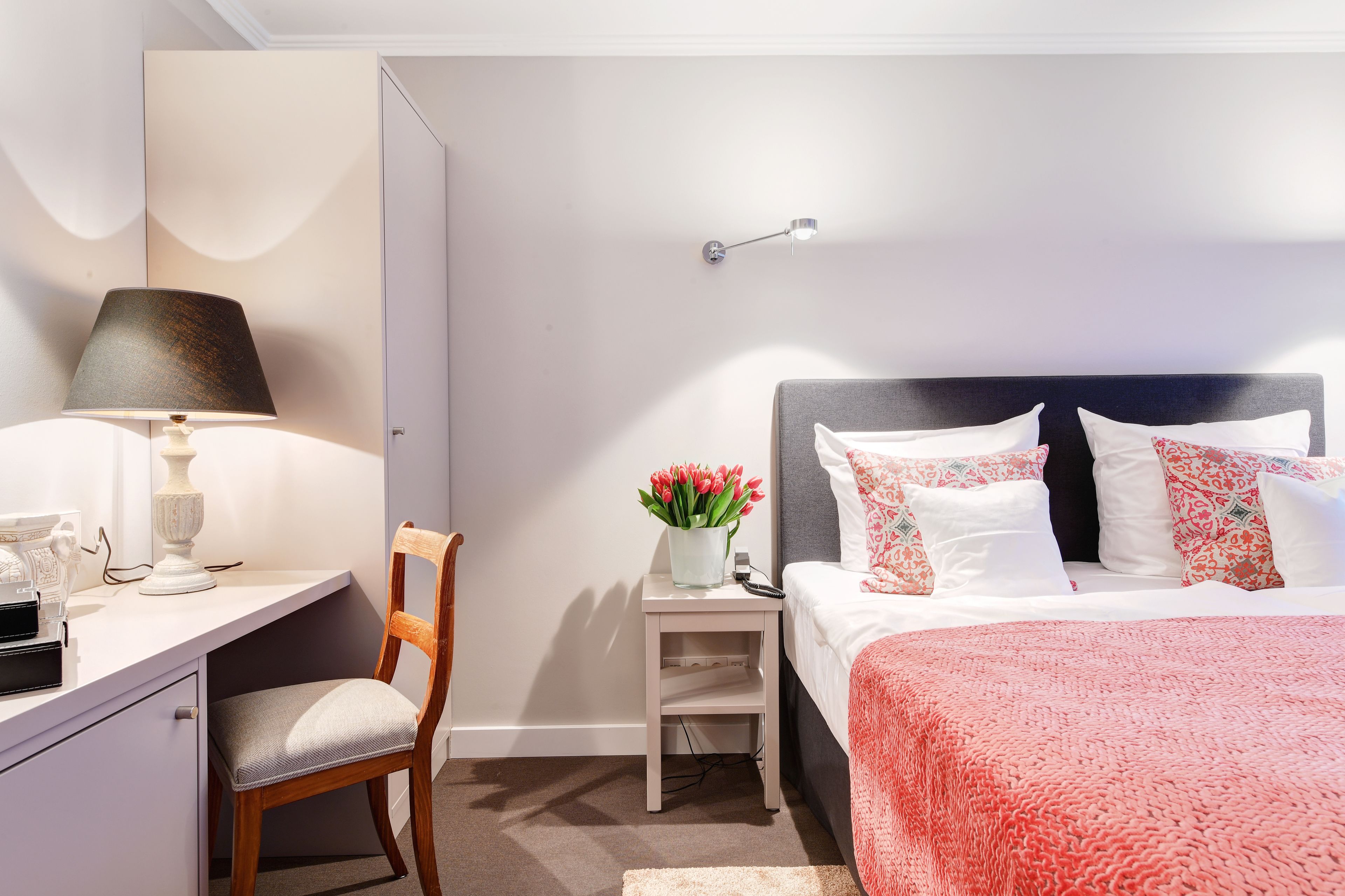 Ein ordentlich eingerichtetes Schlafzimmer mit Bett, Nachttisch, Lampe, Stuhl und Schreibtisch mit Lampe und Blumenvase.