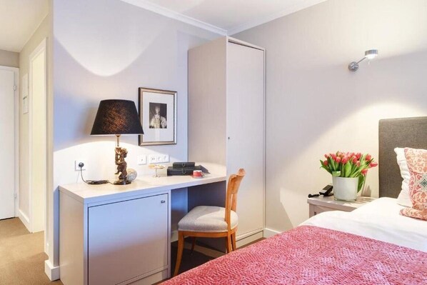 Habitación de hotel pequeña con cama, escritorio con lámpara, silla, armario y mesita de noche con un jarrón de flores.