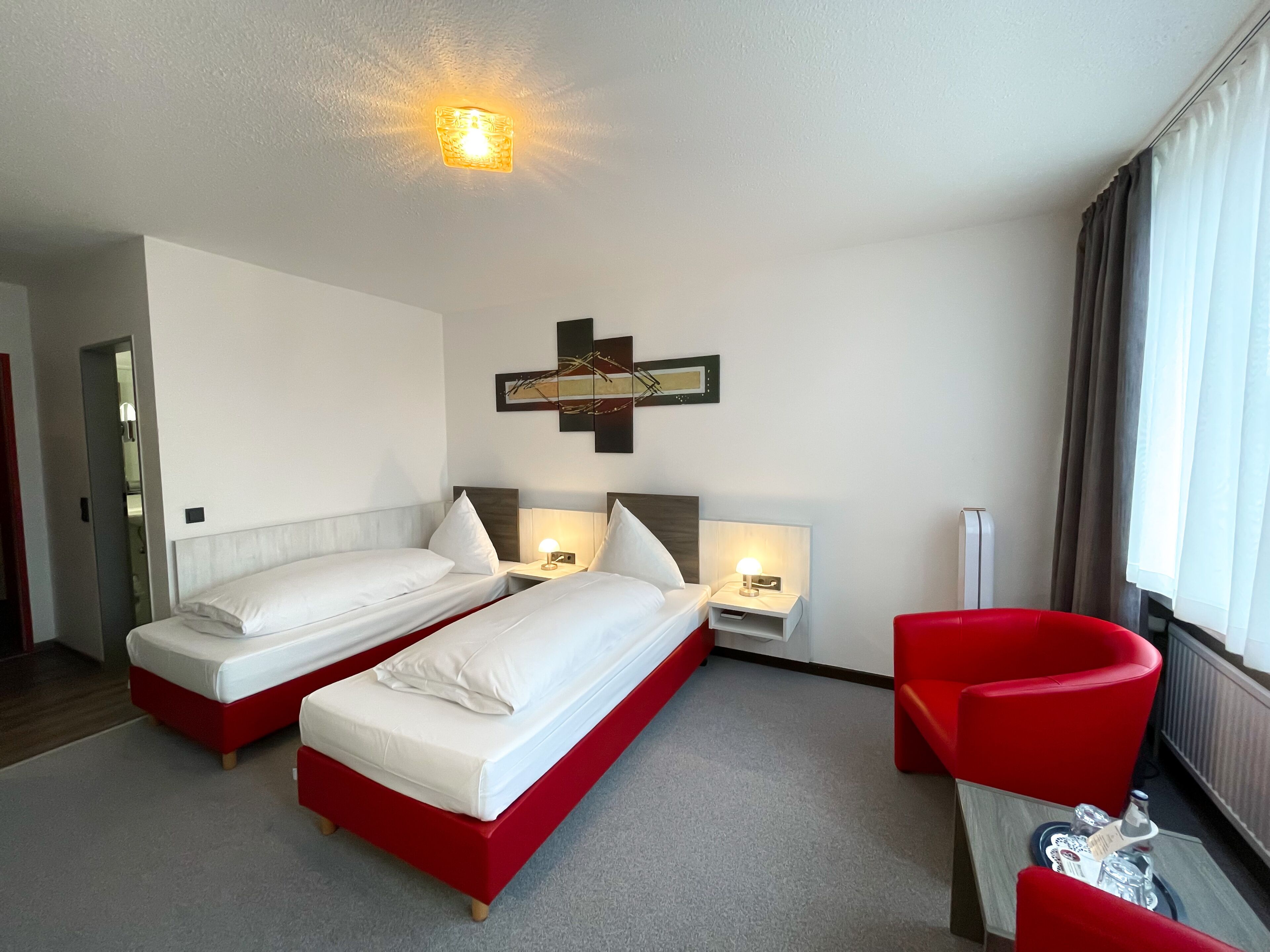 A hotel room with two beds, a red armchair, a small table with glasses, and a ceiling light.