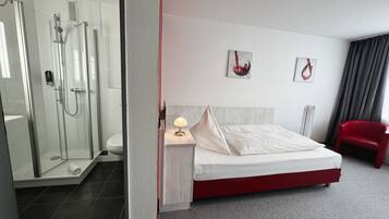 A modern hotel room with a glass-enclosed shower, a neatly made bed, a bedside table with a lamp, and two artwork pieces on the wall.