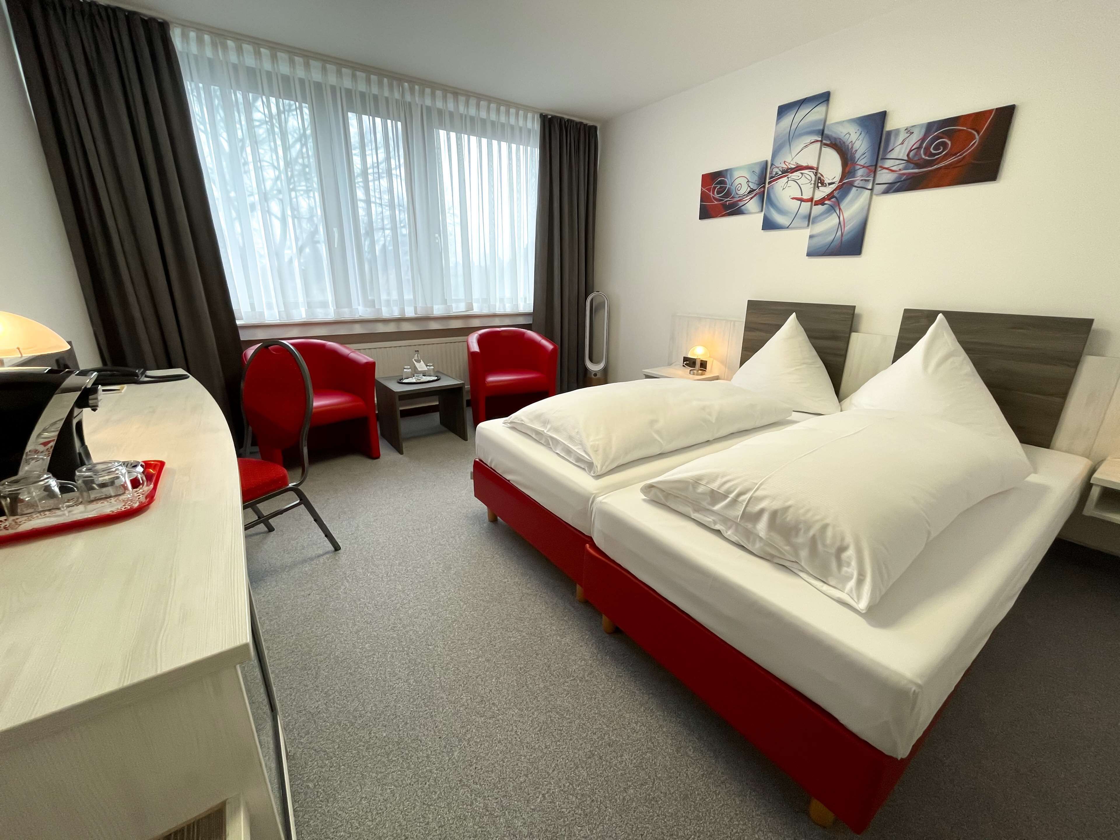 A hotel room with a bed, two red chairs, a desk with a lamp, a coffee machine, and abstract wall art.