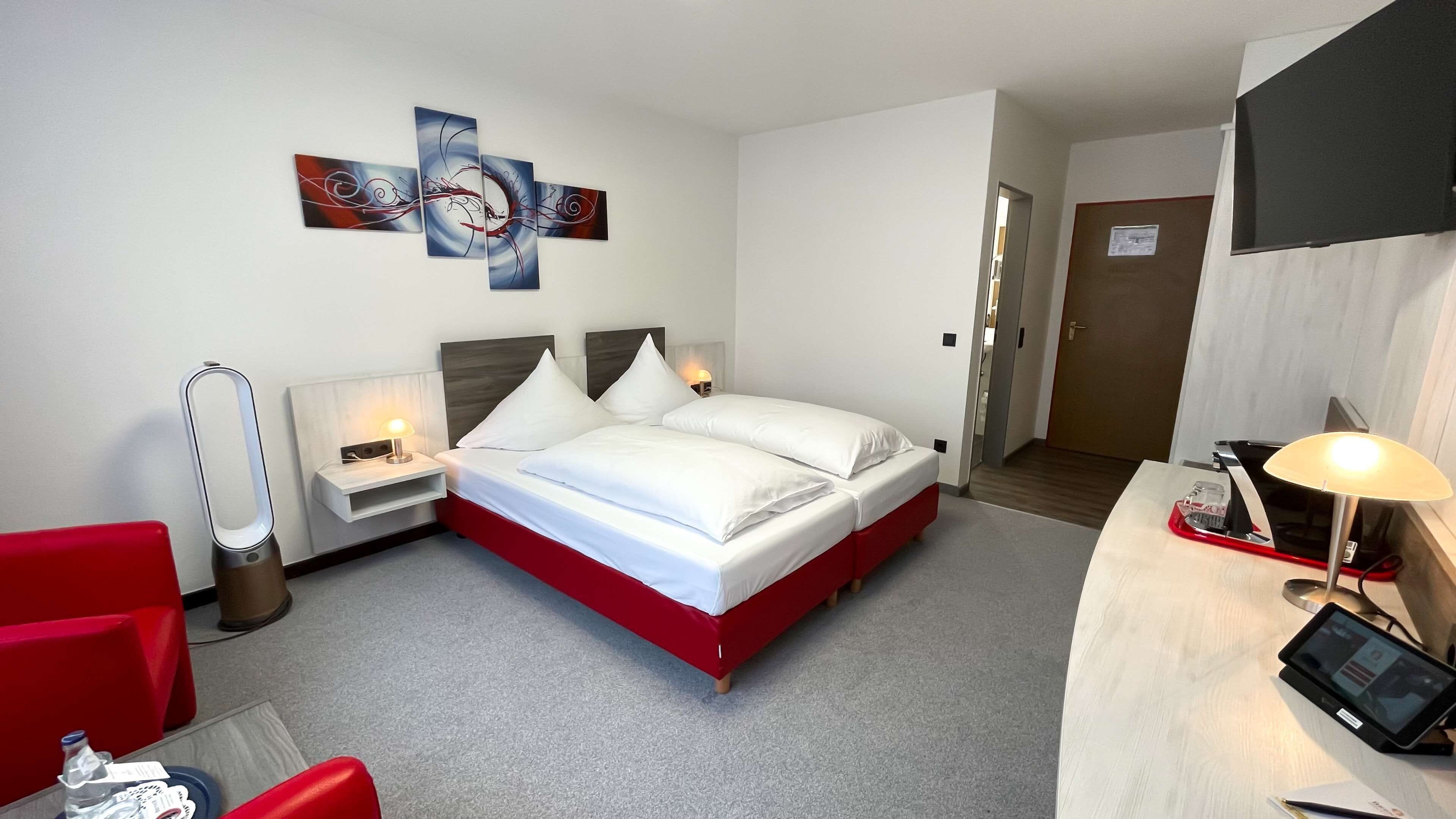 A hotel room with a bed, a desk with a computer, a chair, a bedside table with a lamp, and a wall art piece.
