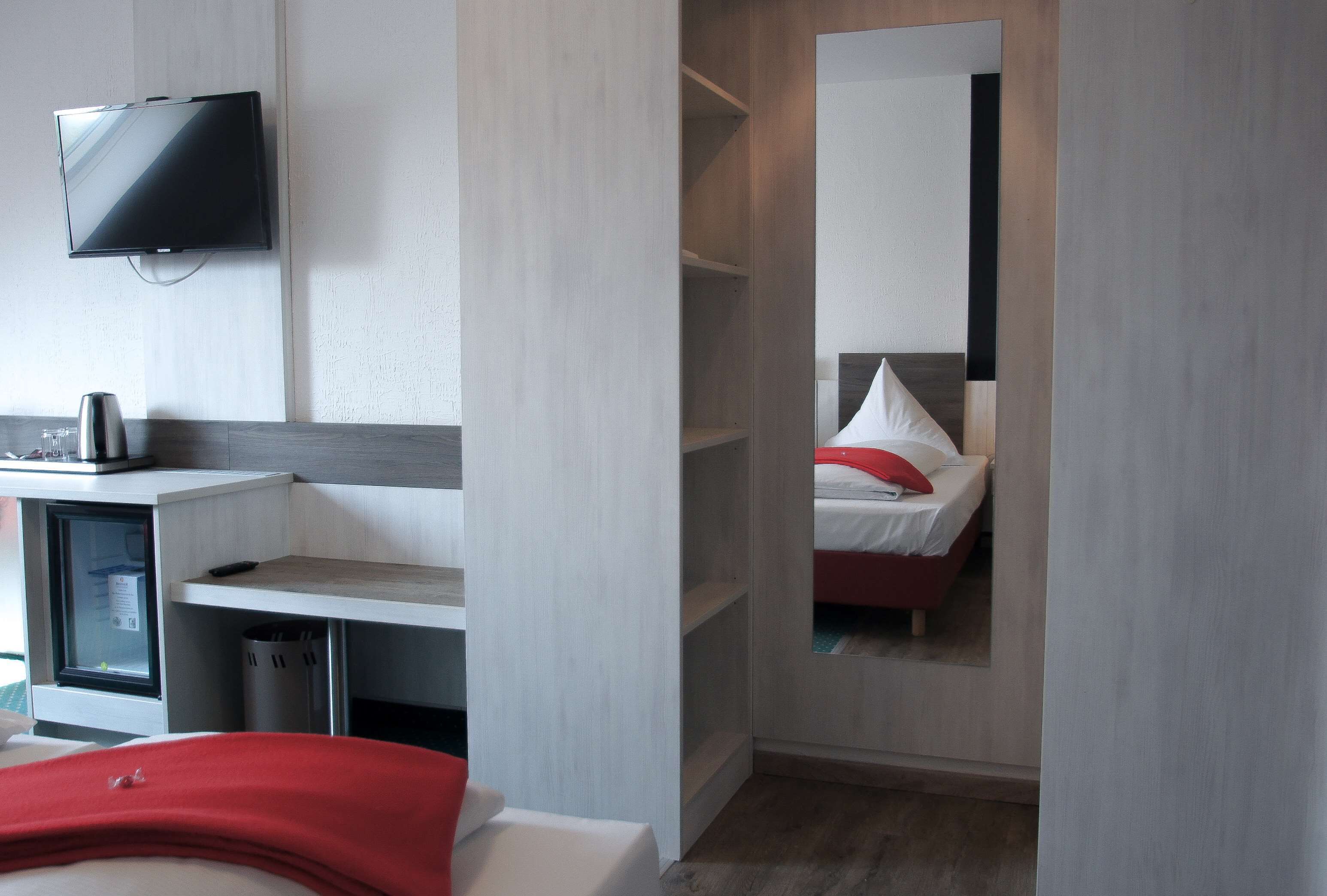 Hotellihuone, jossa on sänky, seinälle asennettu televisio, työpöytä tietokoneella ja peili, joka heijastaa huonetta.