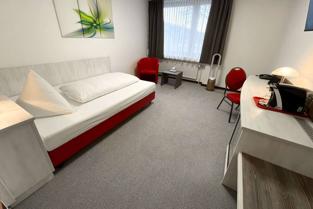 A hotel room with a bed, a desk with a lamp, a chair, a small table, and a radiator.
