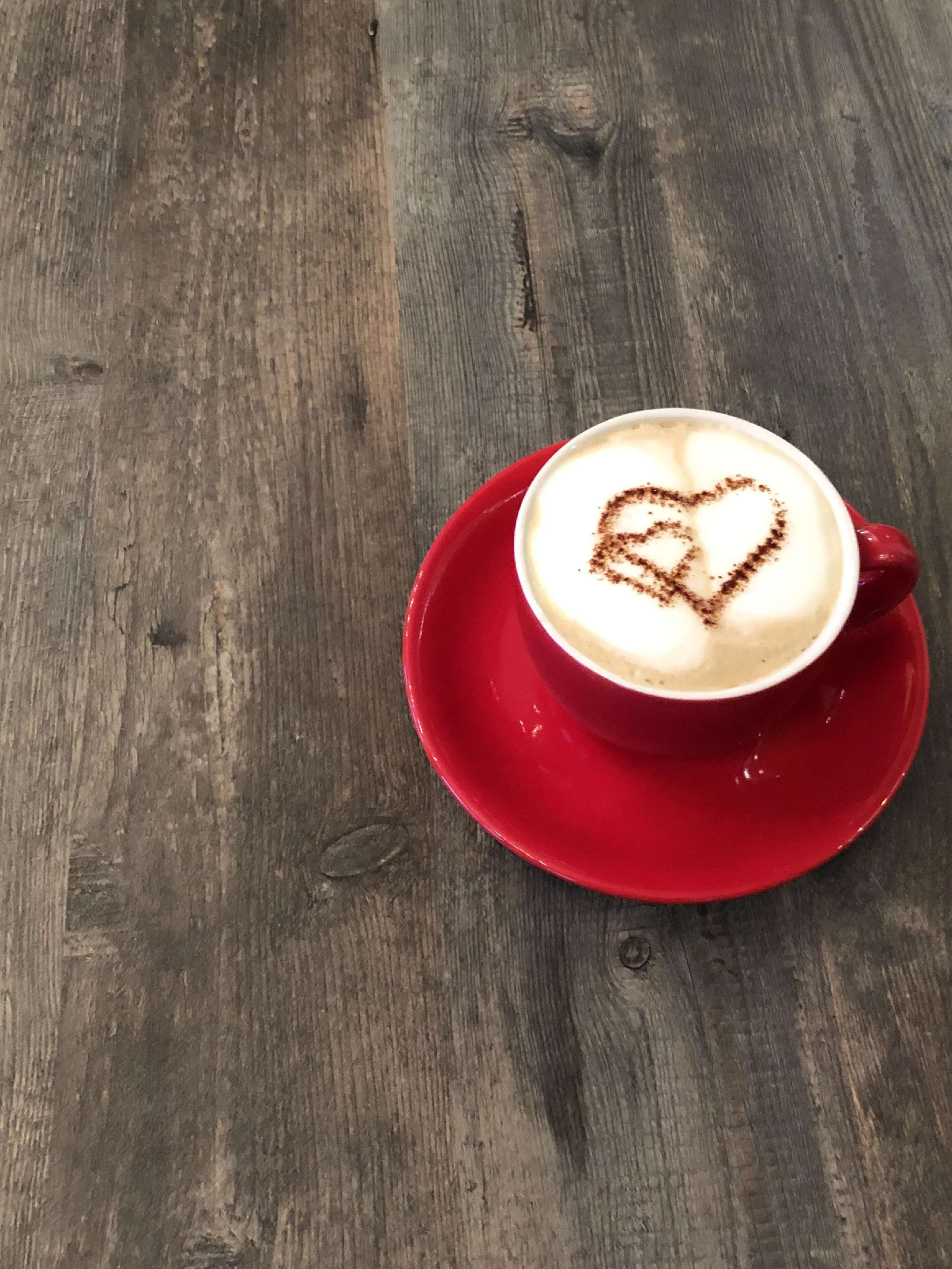 A cup of coffee with a heart-shaped latte art on a red saucer, placed on a wooden table.