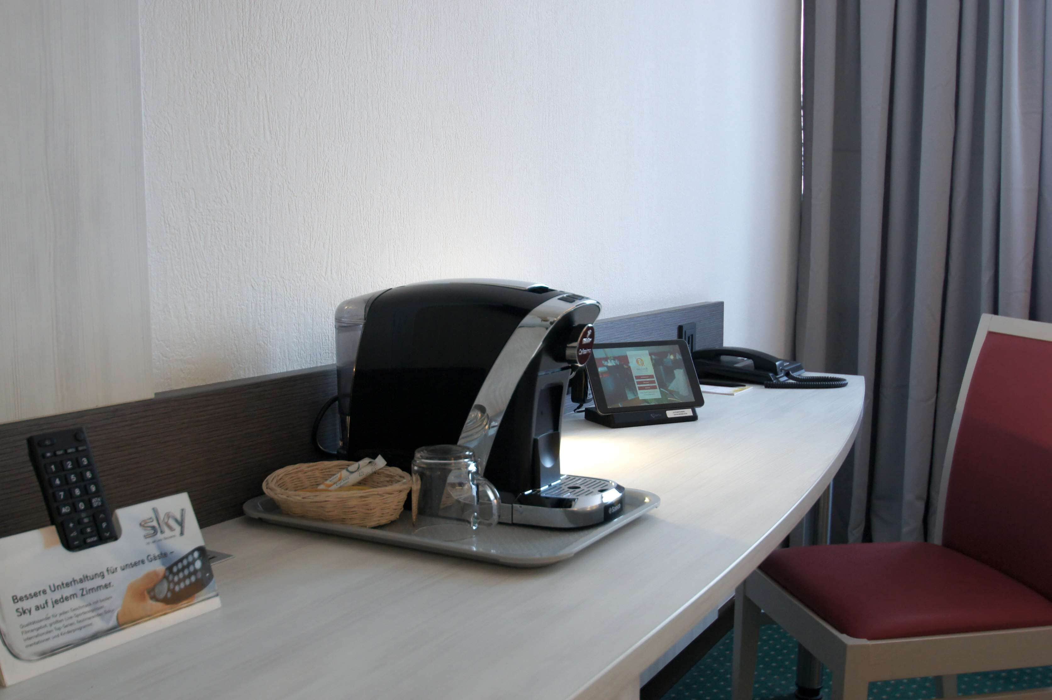 A hotel room with a desk featuring a coffee machine, a remote control, a tablet, and a Sky service sign.