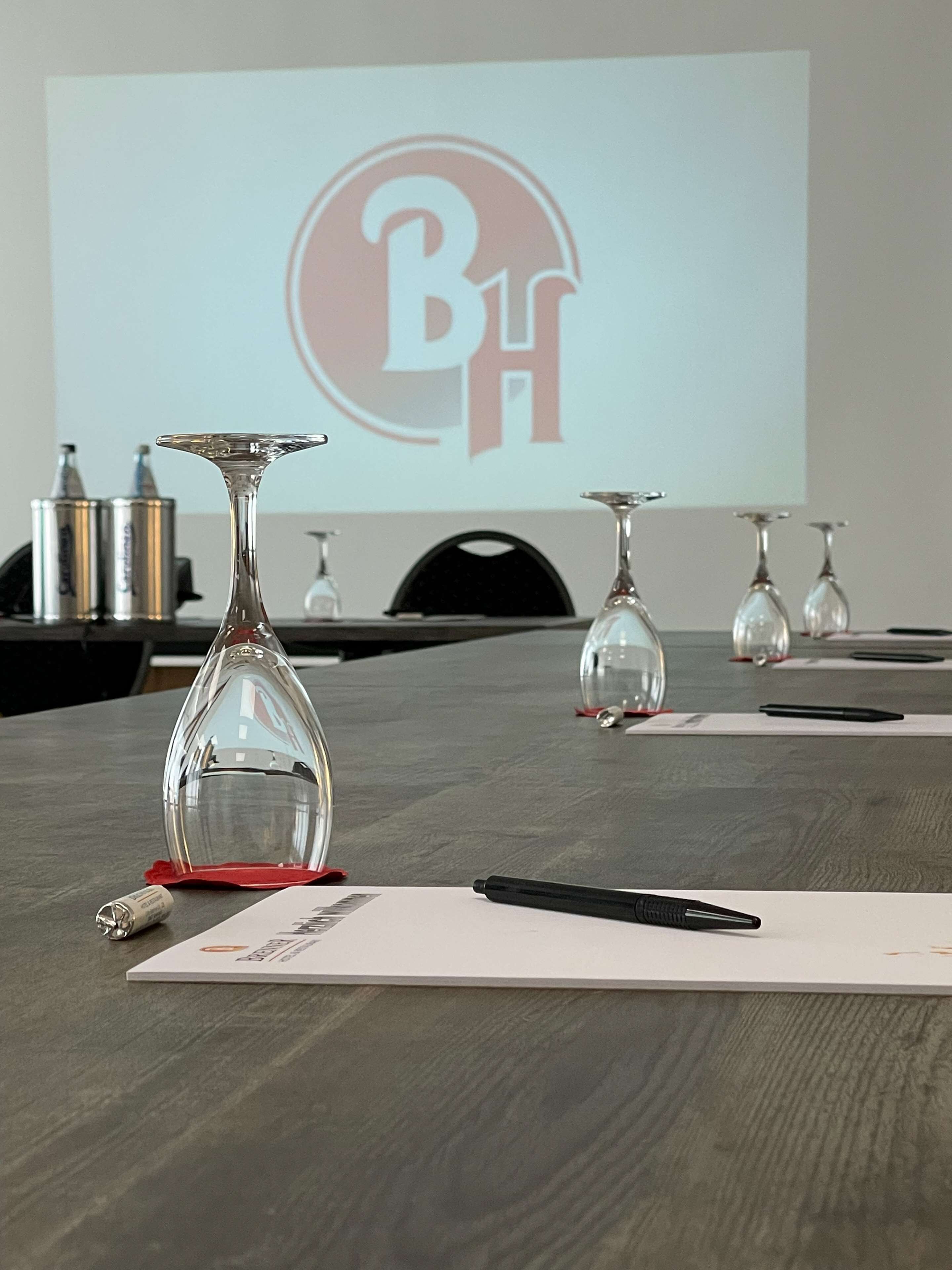Uma sala de conferências com tela de projetor exibindo um logotipo, um vaso de vidro, uma caneta e uma garrafa sobre a mesa.