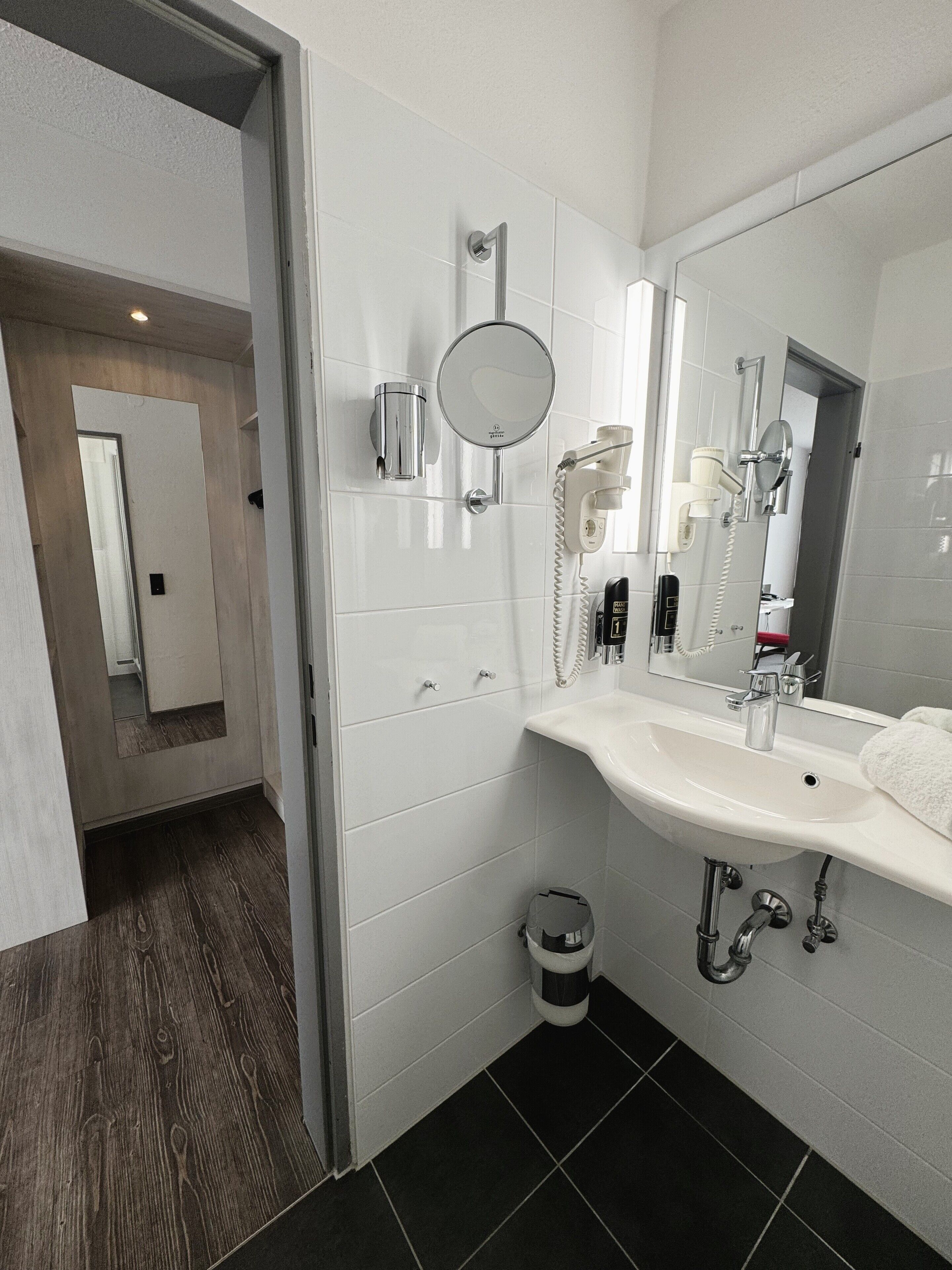 A modern bathroom with a white sink, mirror, and hairdryer. There is a trash can and a shower area visible.