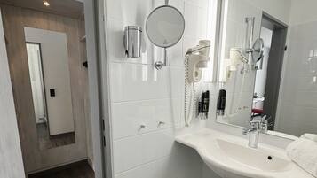 A modern bathroom with a white sink, mirror, and hairdryer. There is a trash can and a shower area visible.
