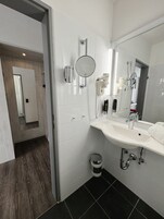 Casa de banho moderna com lavatório branco, espelho e secador de cabelo. É possível ver também uma lixeira e a área do chuveiro.