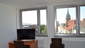 Ein Zimmer mit Schreibtisch, Stuhl und Fernseher. Zwei Fenster bieten Blick auf Gebäude und einen Kirchturm.