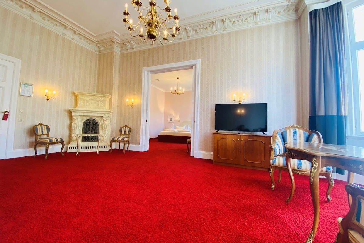 A spacious room with a red carpet, a fireplace, a television, and a large window with curtains.