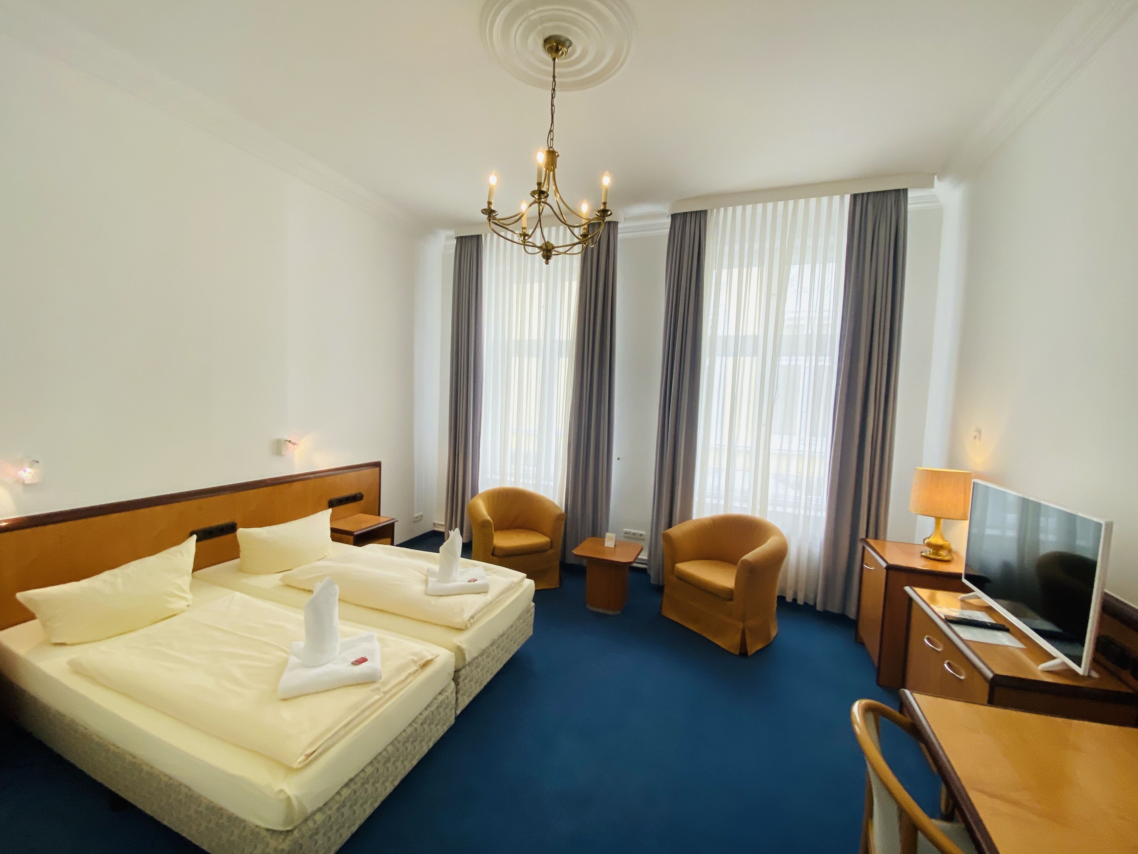 Quarto de hotel com cama de casal, duas cadeiras, escrivaninha com computador, televisão e lustre.