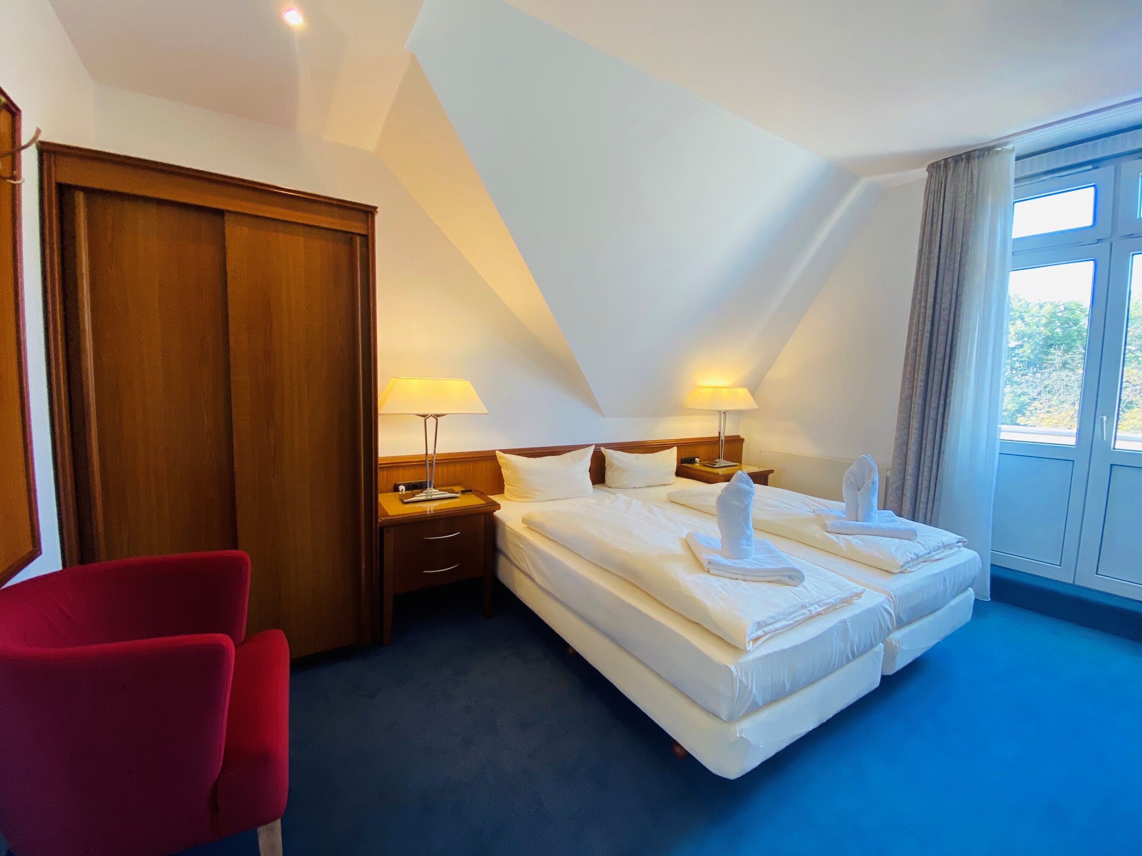 A hotel room with two beds, a red chair, wooden wardrobe, nightstands with lamps, and a large window with curtains.