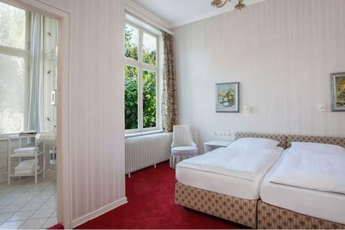 A hotel room with a large bed, a radiator, a chair, and a window with curtains.