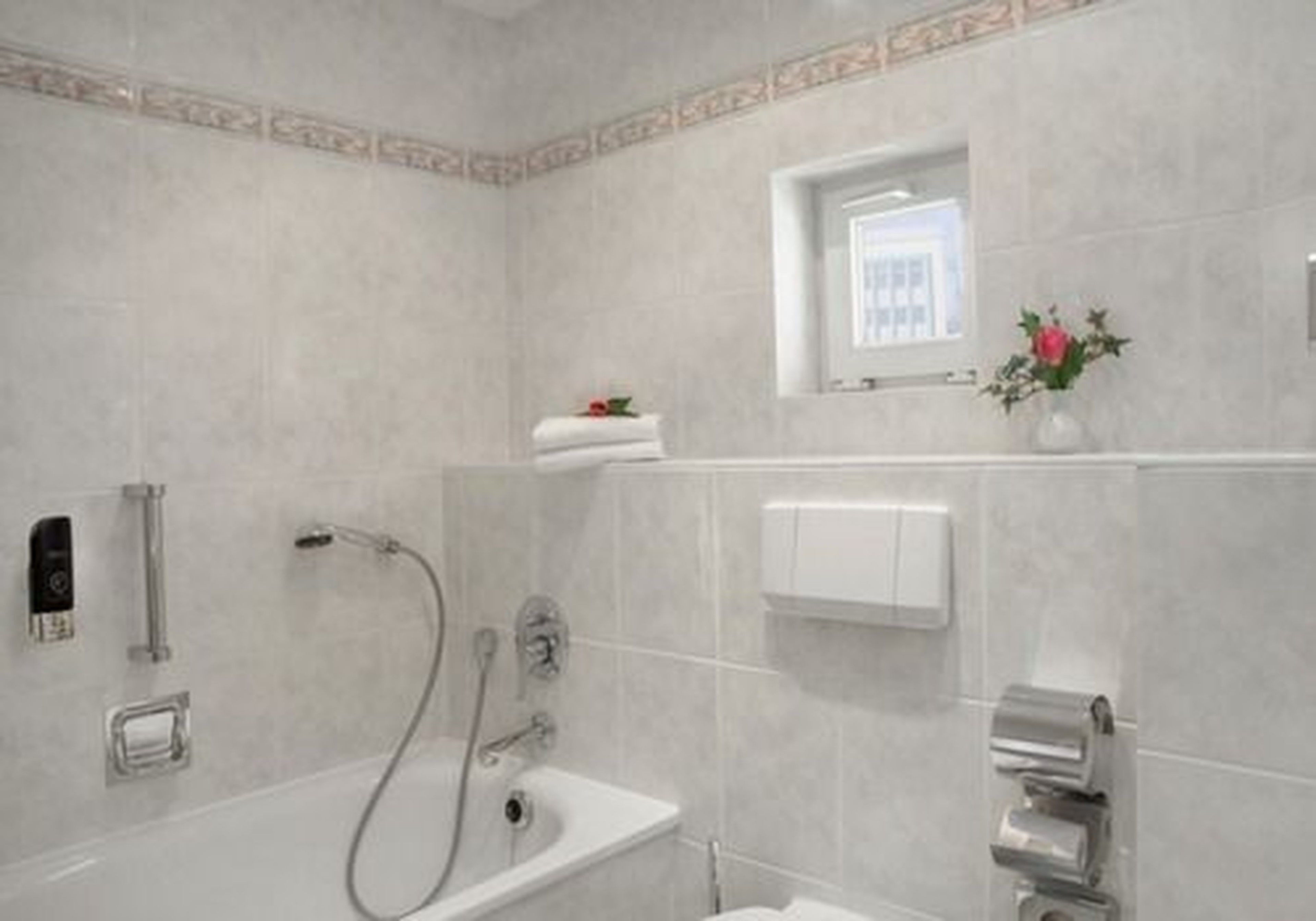 Een moderne badkamer met badkuip, douche en toilet. Er is een raam met uitzicht, een handdoekhouder en een klein plankje met een bloevas.