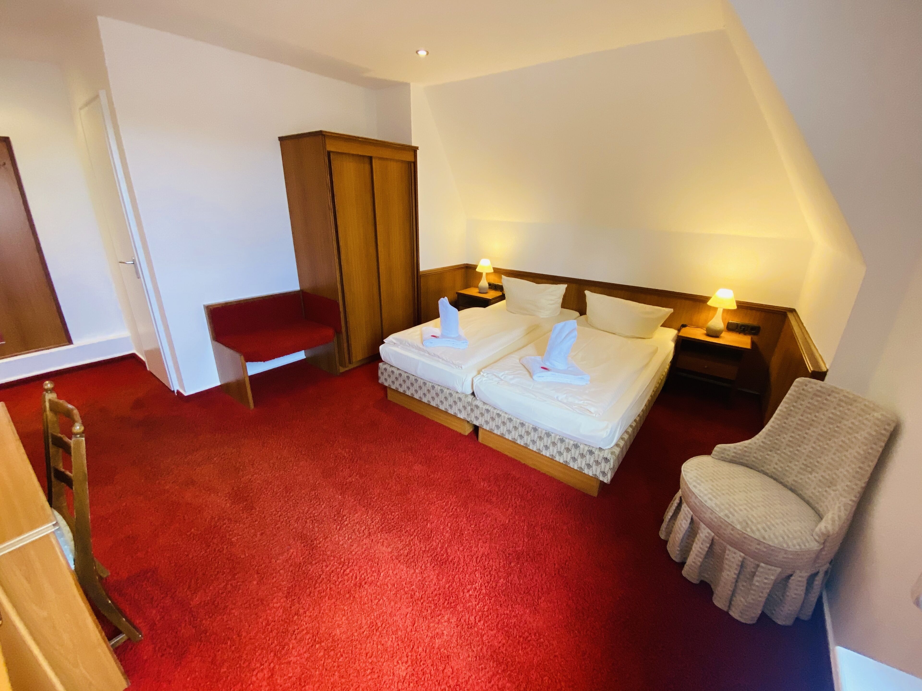 Habitación de hotel con alfombra roja, dos camas con ropa blanca, un armario de madera, una silla roja y dos lámparas de noche.