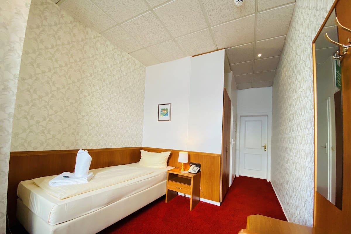 A hotel room with a single bed, a bedside table with a lamp, a phone, and a small drawer.