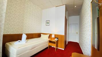 A hotel room with a single bed, a bedside table with a lamp, a phone, and a small drawer.