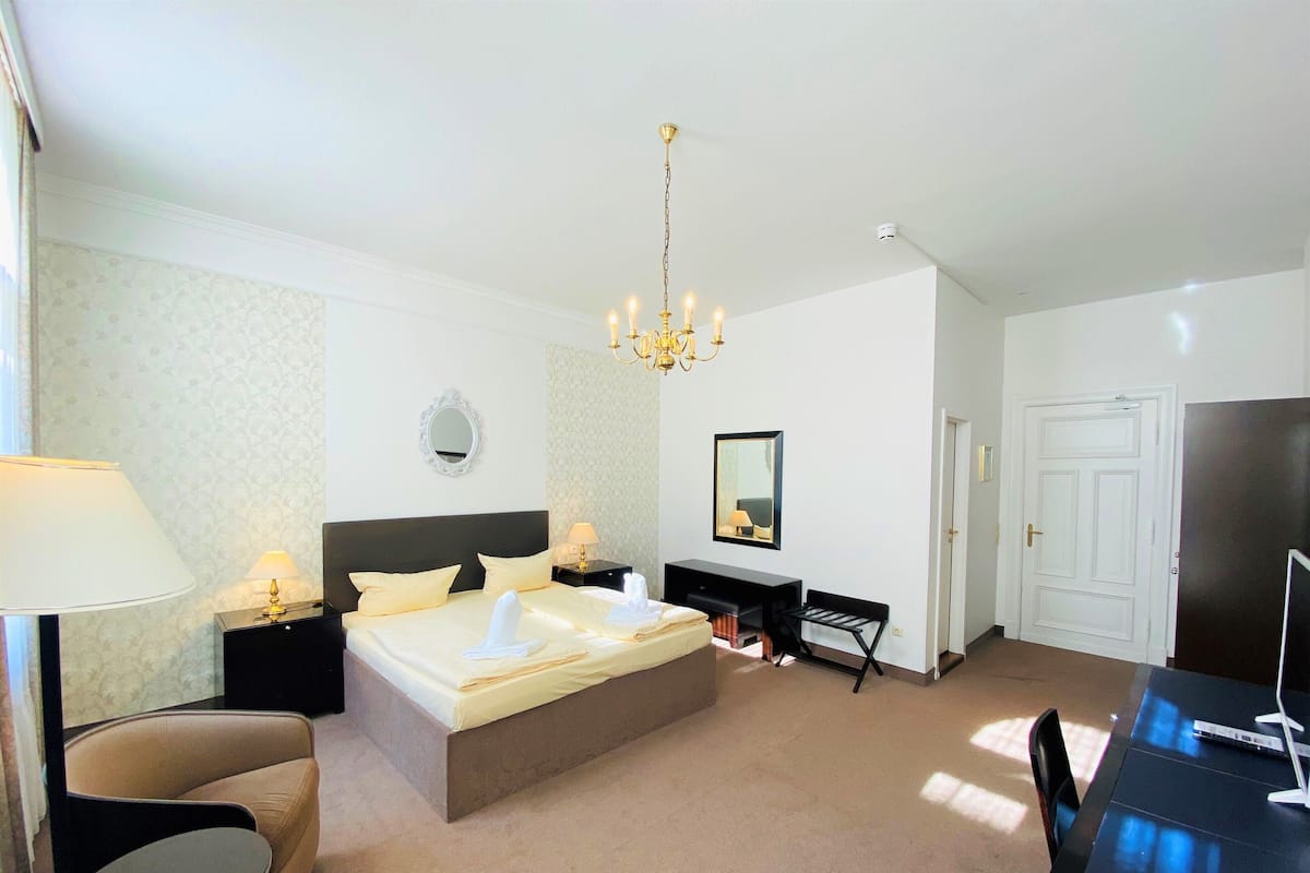 A hotel room with a large bed, a desk, a chair, a mirror, and a chandelier.