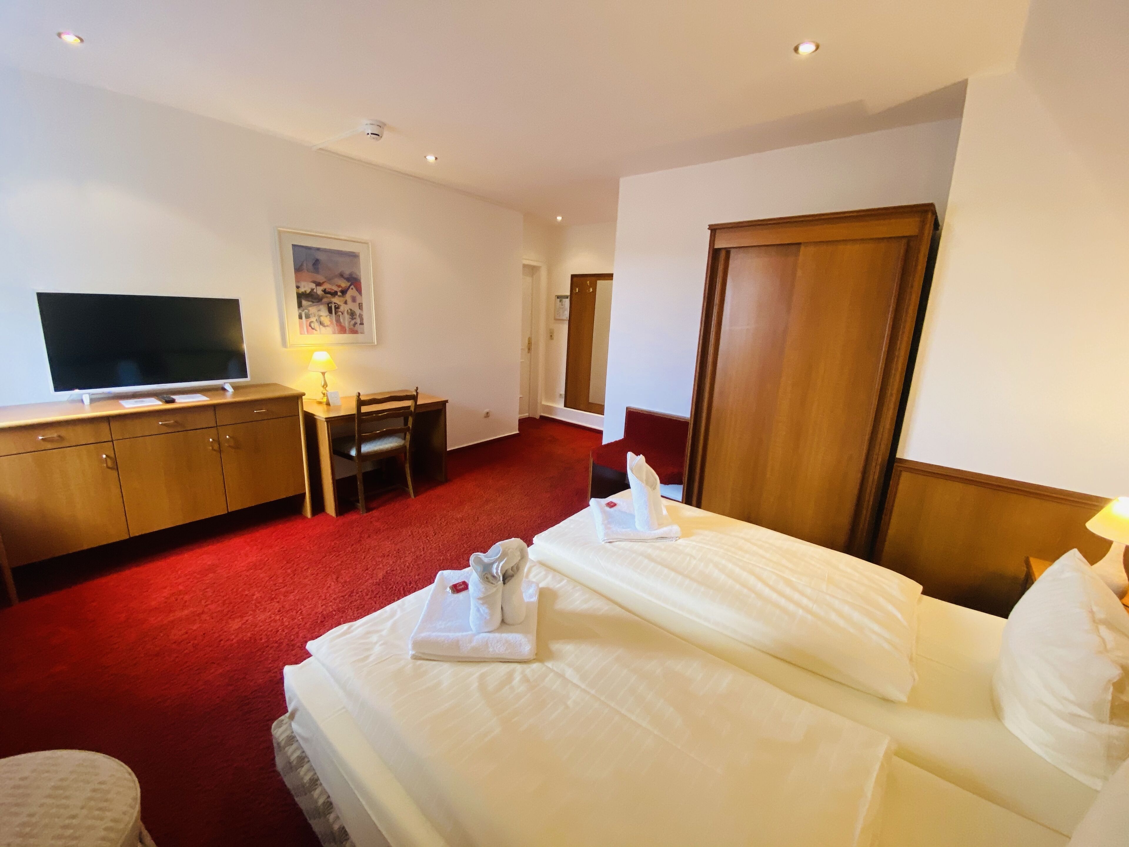 Une chambre d’hôtel avec un grand lit, un bureau avec une chaise, une télévision et une moquette rouge.