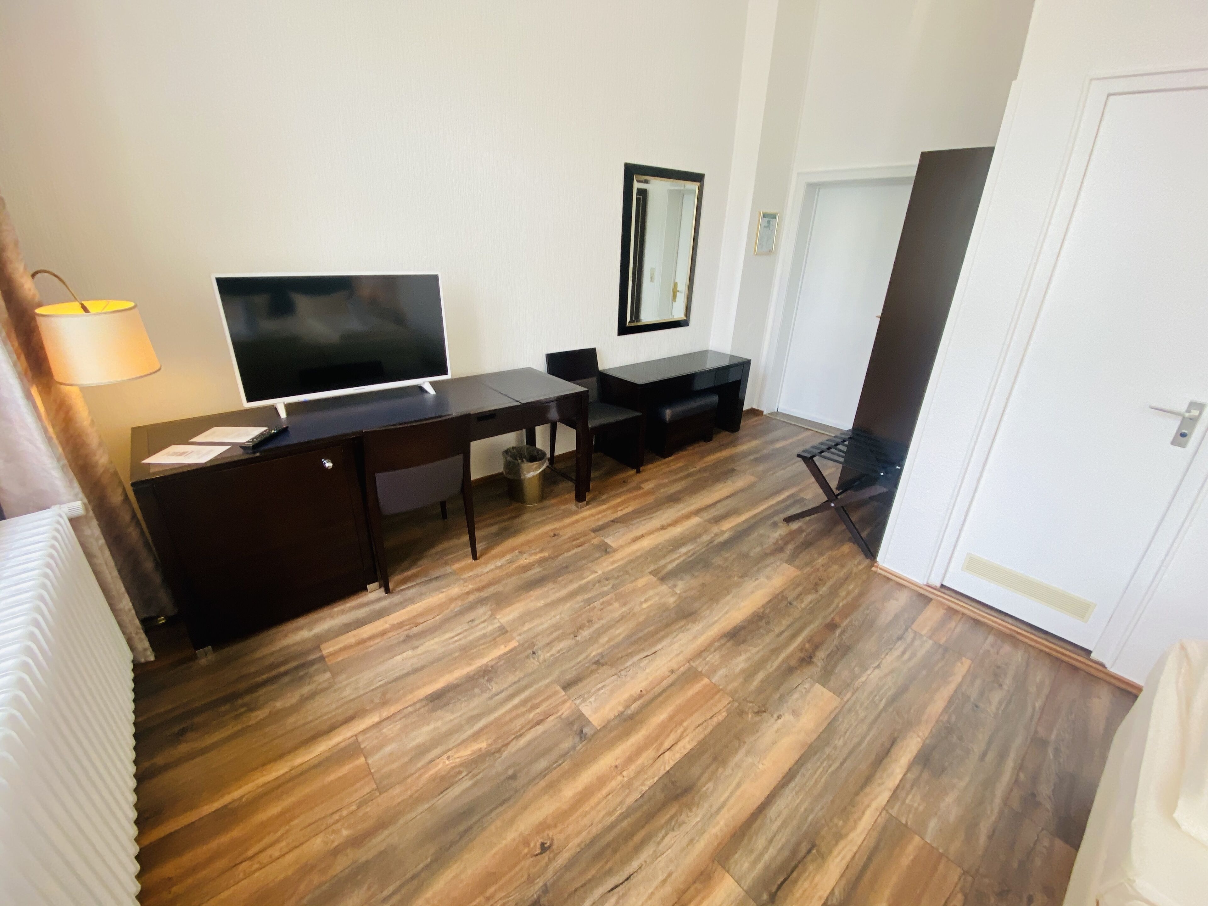 A hotel room with a wooden floor, a desk with a flat-screen TV, a chair, a small table, and a lamp.