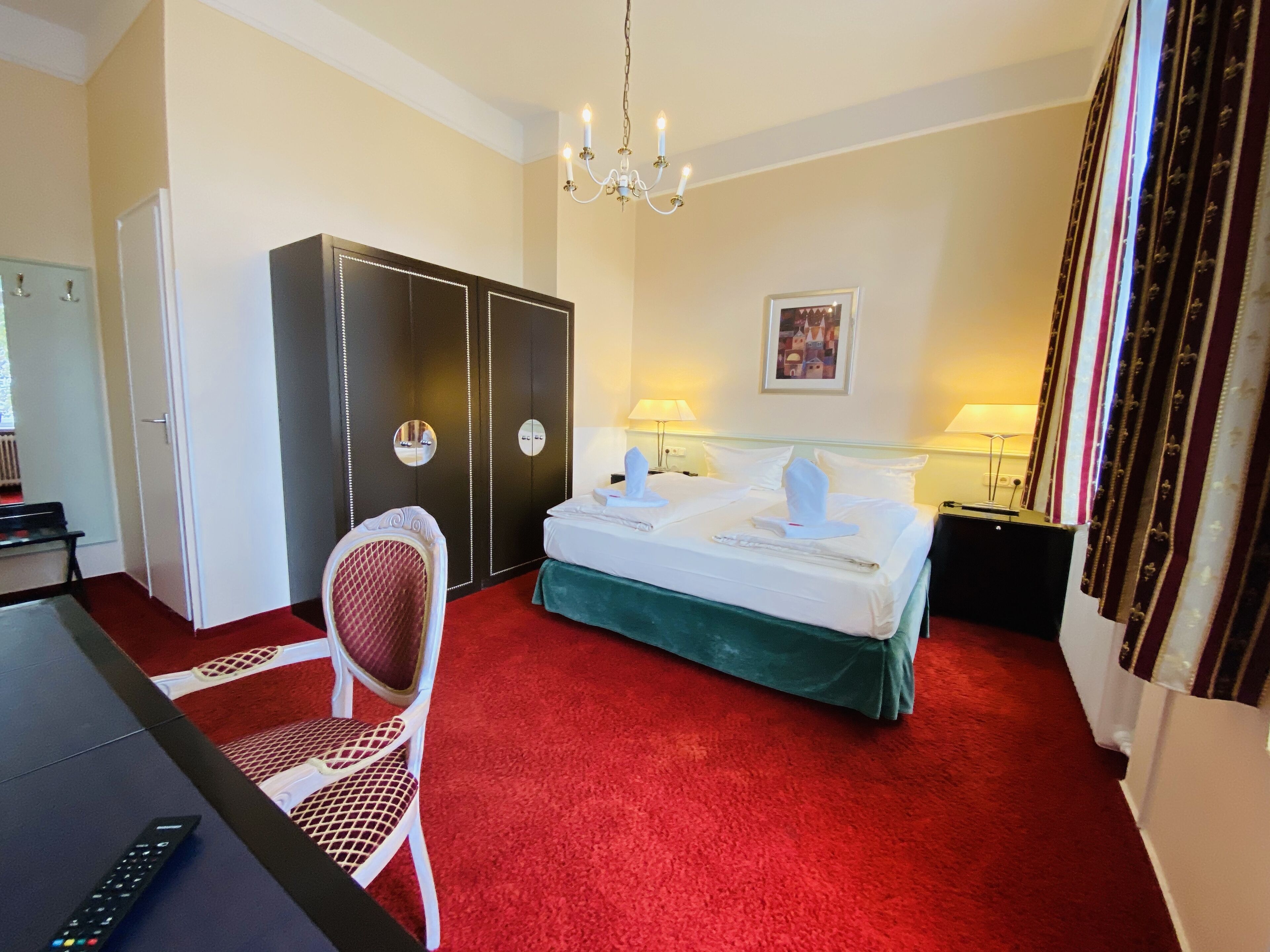 A hotel room with a bed, a desk, a chair, a wardrobe, and a chandelier.