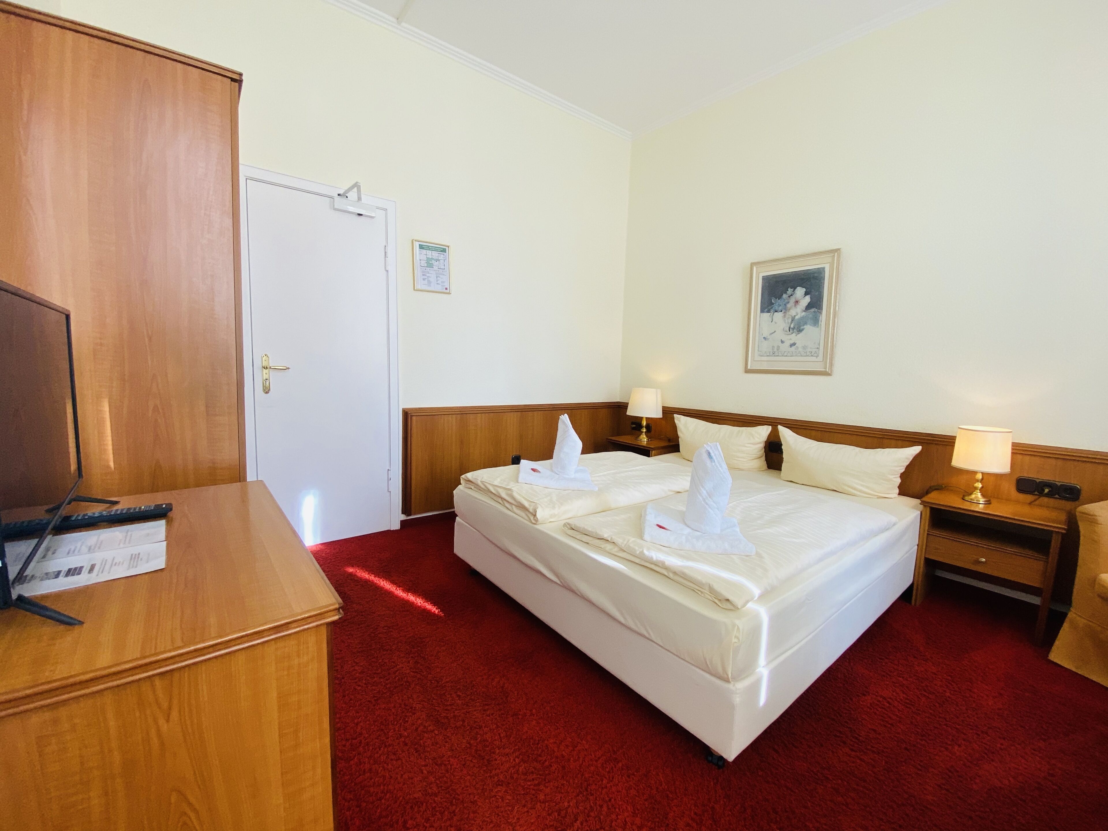 A hotel room with a wooden wardrobe, a desk with a keyboard and mouse, a bed with white linens and towels, a nightstand with a lamp, and a framed picture on the wall.