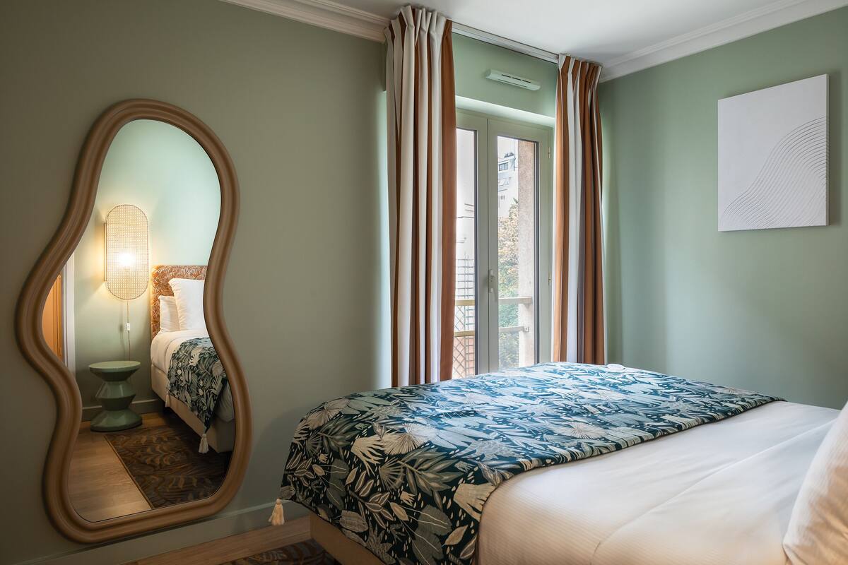 Une chambre à coucher comprenant un lit, un miroir, une fenêtre avec des rideaux et une œuvre d’art accrochée au mur.