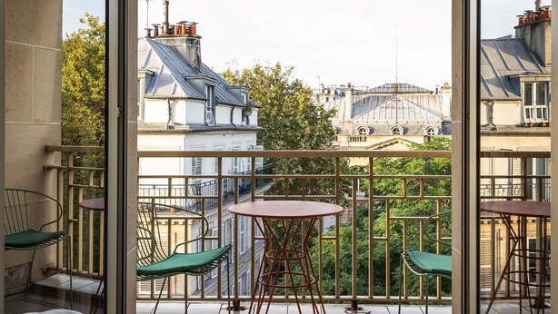 Une chambre avec un grand lit, un balcon avec des chaises et une table, et une vue sur les bâtiments et les arbres.
