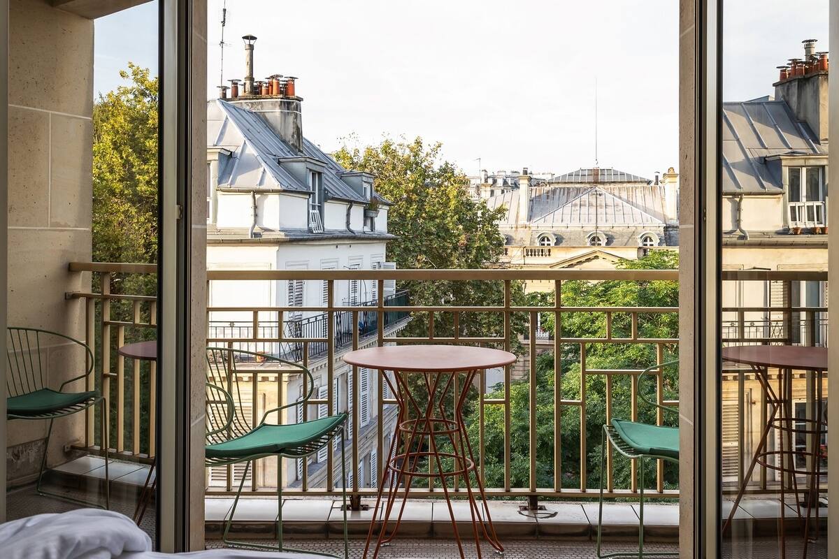 Une chambre avec un grand lit, un balcon avec des chaises et une table, et une vue sur les bâtiments et les arbres.