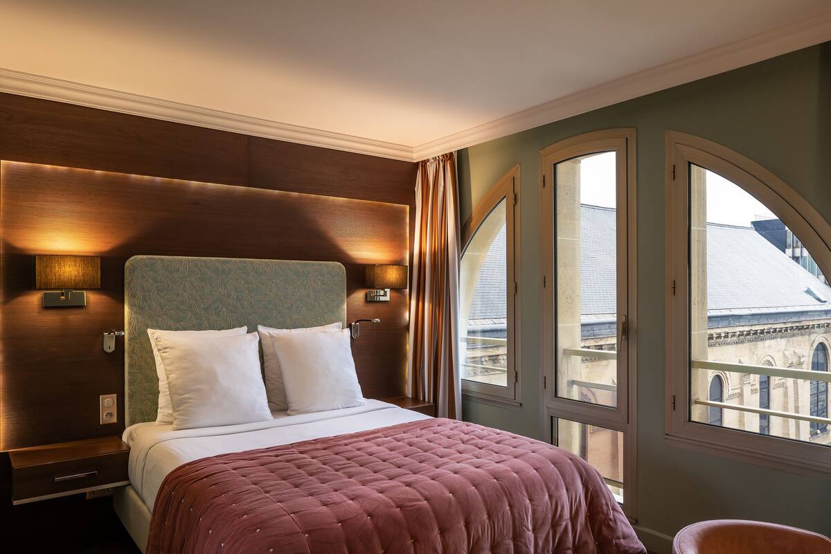 Une chambre d’hôtel avec un grand lit, une tête de lit en bois, deux lampes de chevet et une vue sur un bâtiment à travers des fenêtres en arc de cercle.