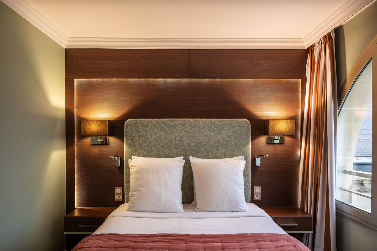 Une chambre d’hôtel avec un grand lit, deux lampes de chevet, une tête de lit en bois et une fenêtre avec des rideaux.