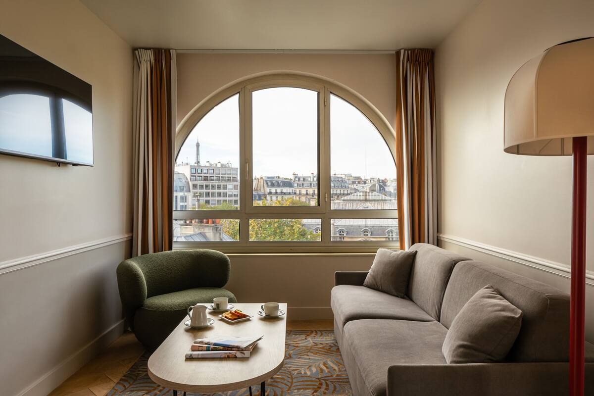 Une chambre d’hôtel moderne dotée d’une grande fenêtre en arc de cercle, d’un canapé, d’un fauteuil vert, d’une petite table sur laquelle se trouve un plateau avec des articles pour le petit-déjeuner, et d’un téléviseur à écran plat fixé au mur.