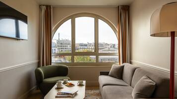 Une chambre d’hôtel avec une grande fenêtre en arc de cercle, un canapé, un fauteuil, une petite table avec un journal et une lampe.