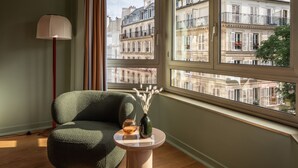 Une pièce avec une grande fenêtre, un fauteuil vert, une petite table ronde ornée d’un vase et d’un verre, et un lampadaire.