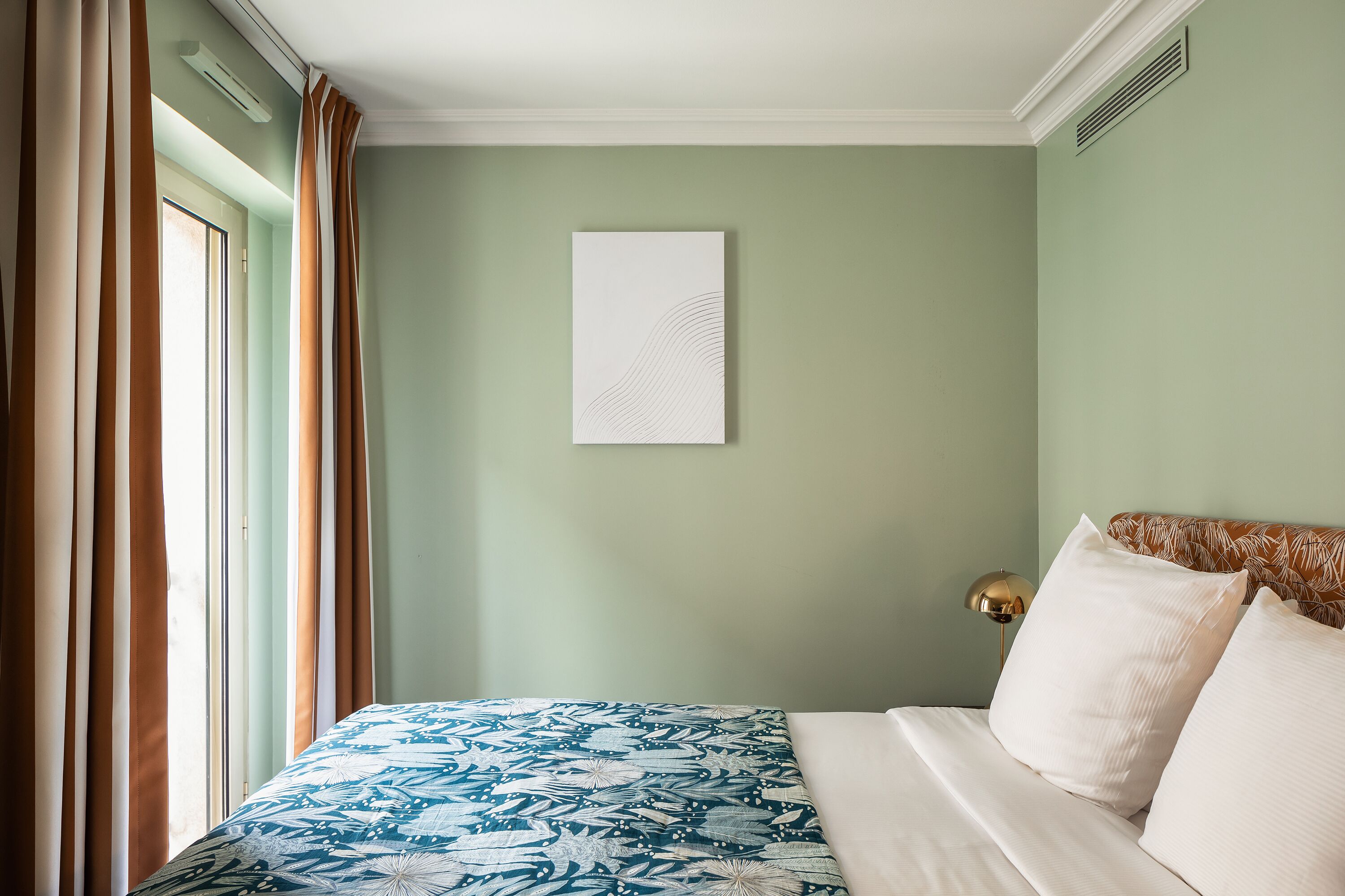 Un lit bien fait, recouvert d’une couvre-lit à motifs, un mur blanc orné d’une œuvre d’art simple, et une fenêtre avec des rideaux.
