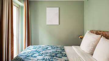 Un lit bien fait, recouvert d’une couvre-lit à motifs, un mur blanc orné d’une œuvre d’art simple, et une fenêtre avec des rideaux.