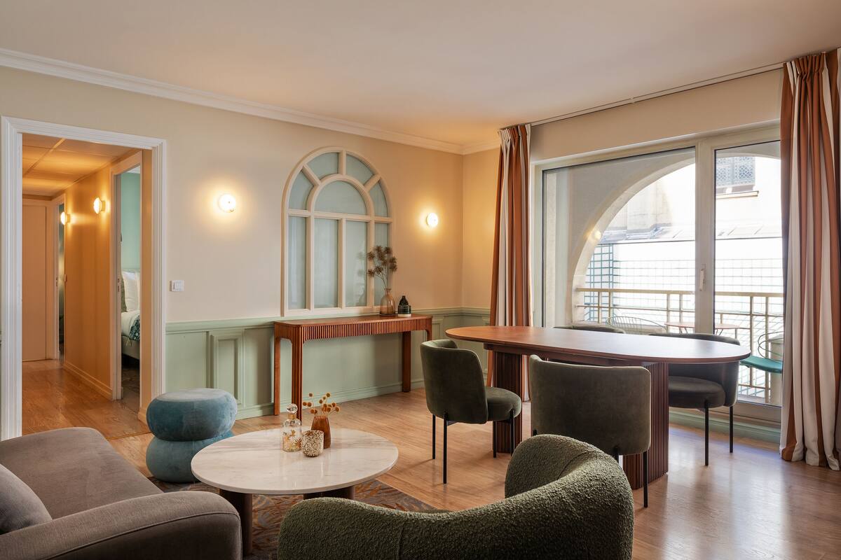 Un salon moderne avec une table de salle à manger ronde, deux fauteuils et un canapé. On y trouve une grande fenêtre avec des rideaux, une porte donnant sur une autre pièce et un luminaire fixé au mur.