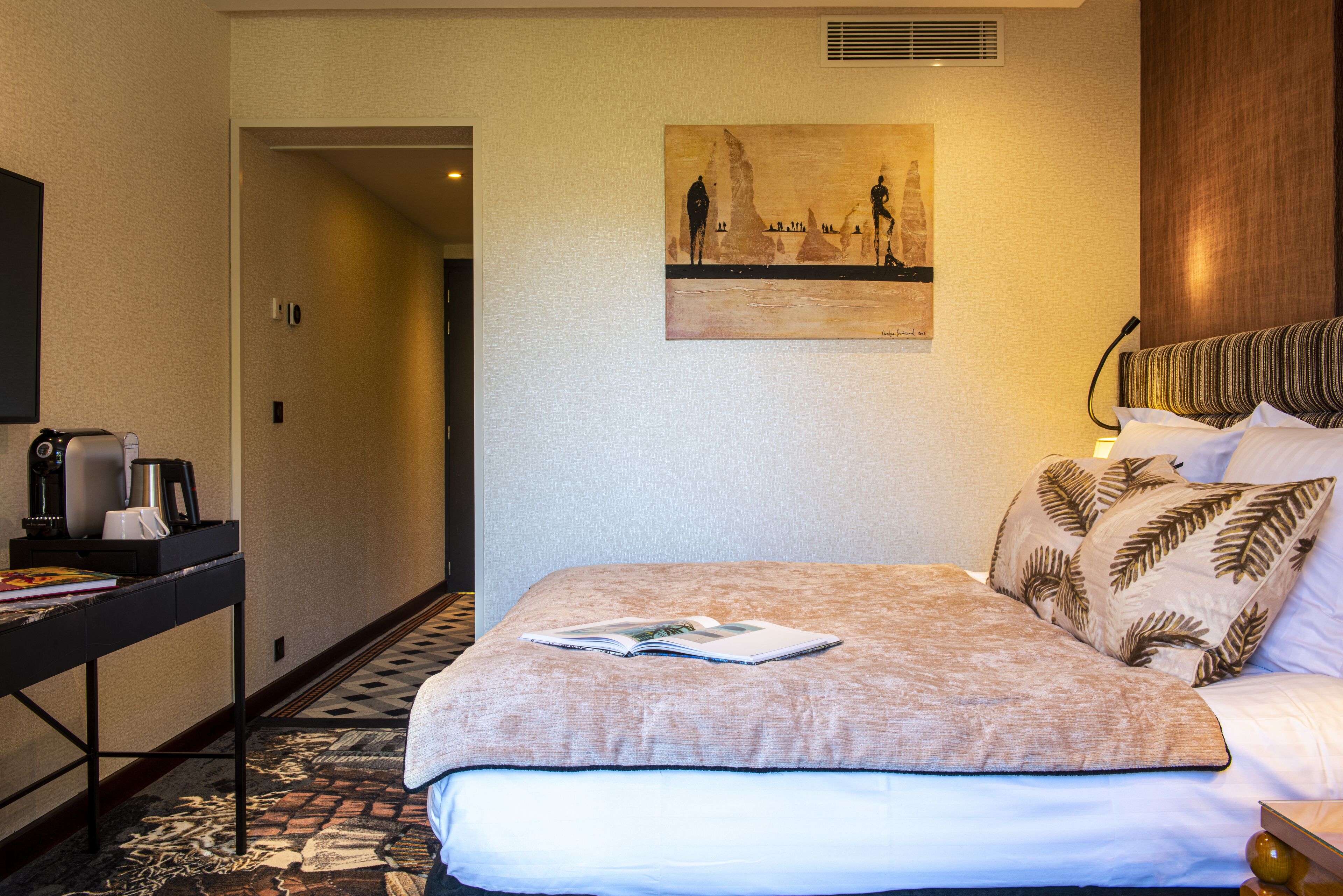 A hotel room with a bed, a painting on the wall, a desk with a coffee maker, and a patterned rug.