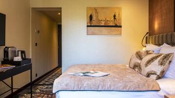 A hotel room with a bed, a painting on the wall, a desk with a coffee maker, and a patterned rug.