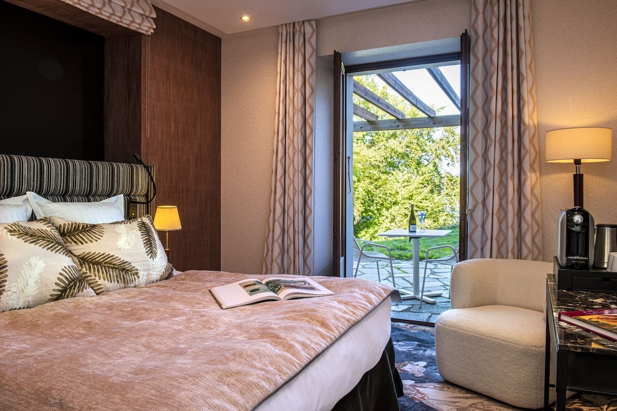 Habitación de hotel con cama, lámpara de lectura, silla, cafetera y vistas al exterior.