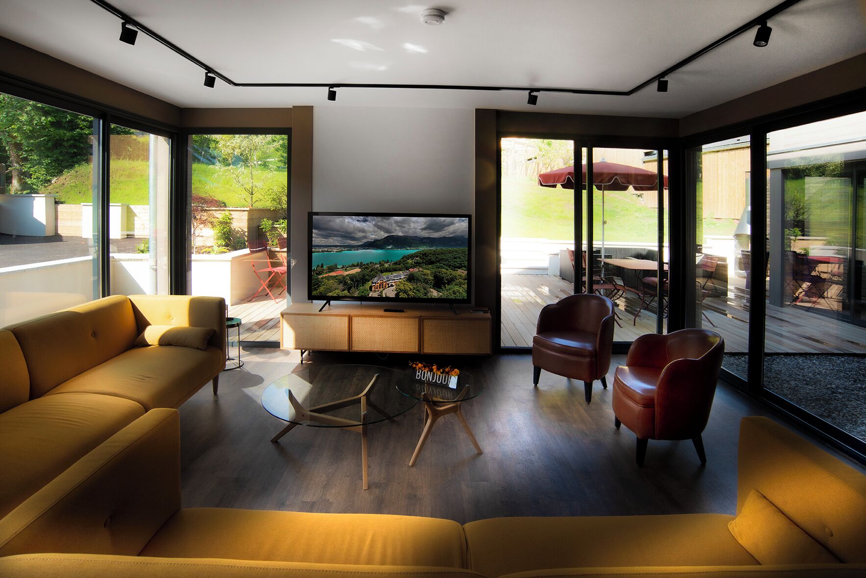 A modern living room with a large flat-screen TV, comfortable seating, and large windows offering a view of the outdoors.