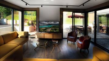 A modern living room with a large flat-screen TV, comfortable seating, and large windows offering a view of the outdoors.
