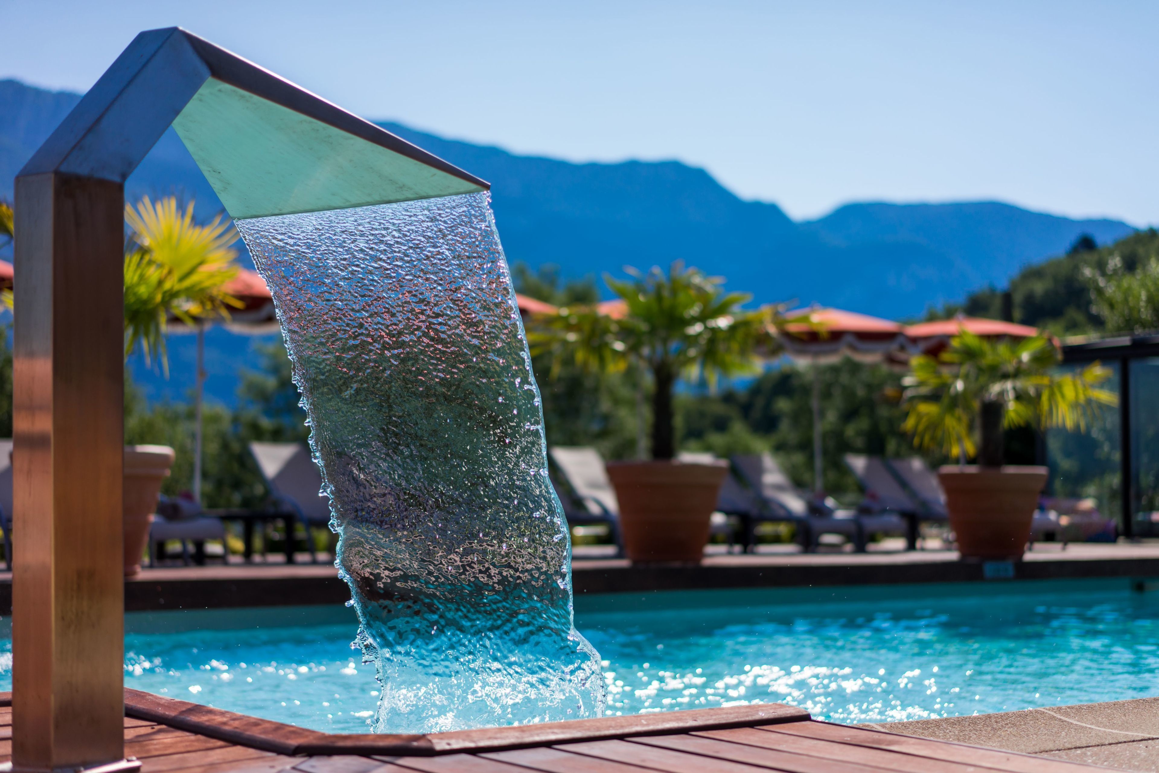 Ein Pool mit Wasserfall, umgeben von Liegestühlen und Topfpflanzen, mit Bergen im Hintergrund.