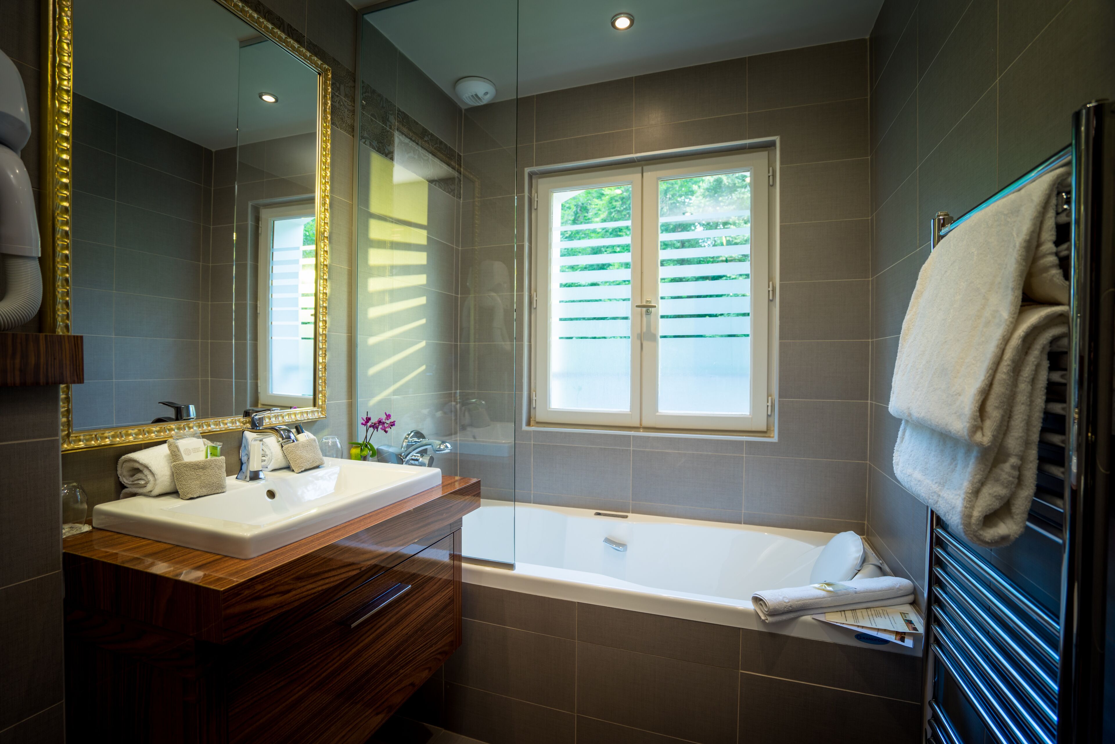 Een moderne badkamer met een houten wastafelmeubel, een grote spiegel, een bad en een raam met zonwering.