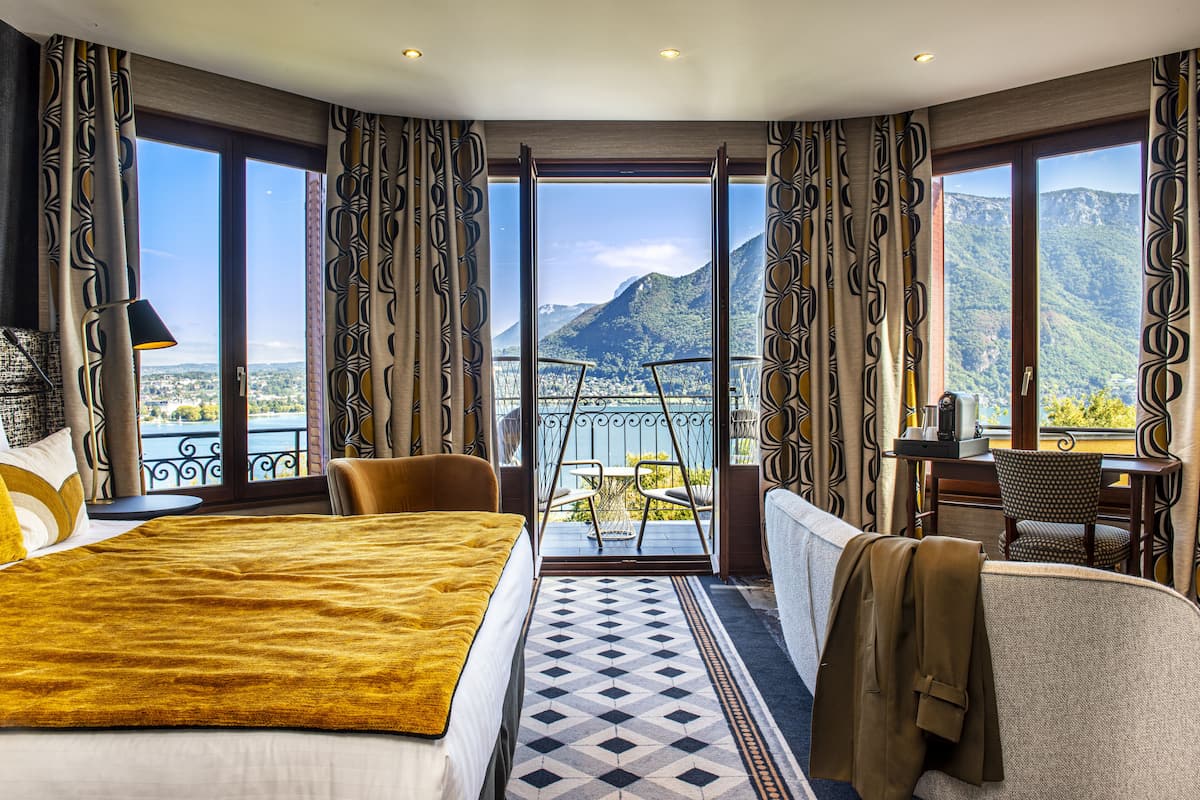 Una habitación de hotel con una cama grande, un sofá, un balcón y vistas a montañas y un lago.