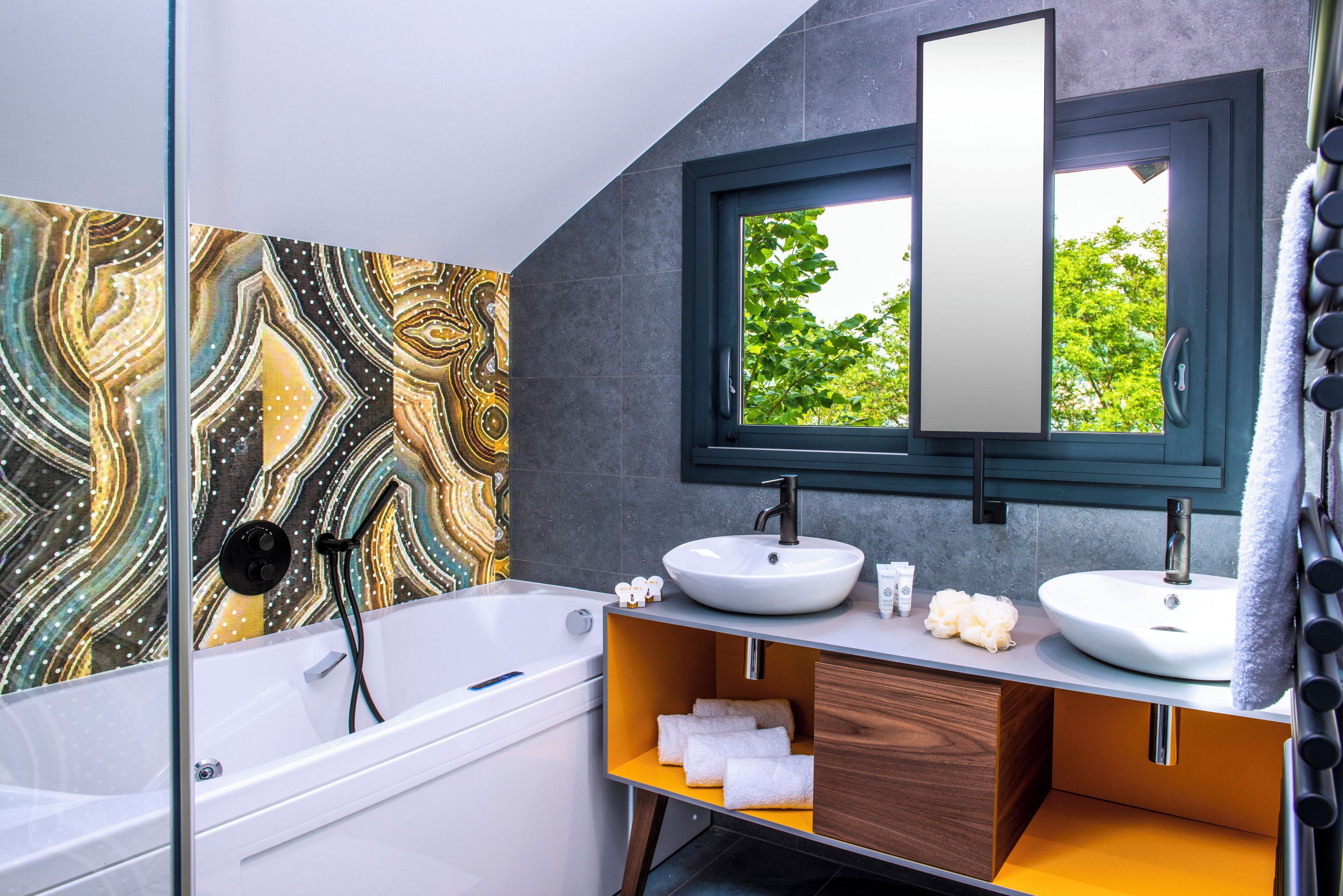Une salle de bain moderne avec une baignoire, deux lavabos, un grand miroir et une vue sur les arbres par la fenêtre.